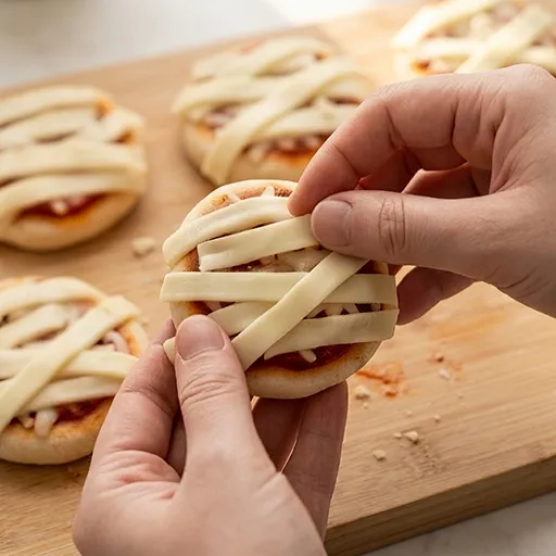 Hands wrapping strips of cheese in a criss-cross pattern over a mini pizza to create the mummy bandage effect