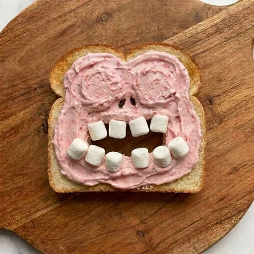 Toast spread with pink cream base, cookie eyes and marshmallow teeth already placed, before adding the pretzel limbs and tongue
